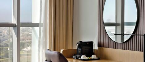 A hotel room with a large window, a desk with a coffee maker, and a mirror.
