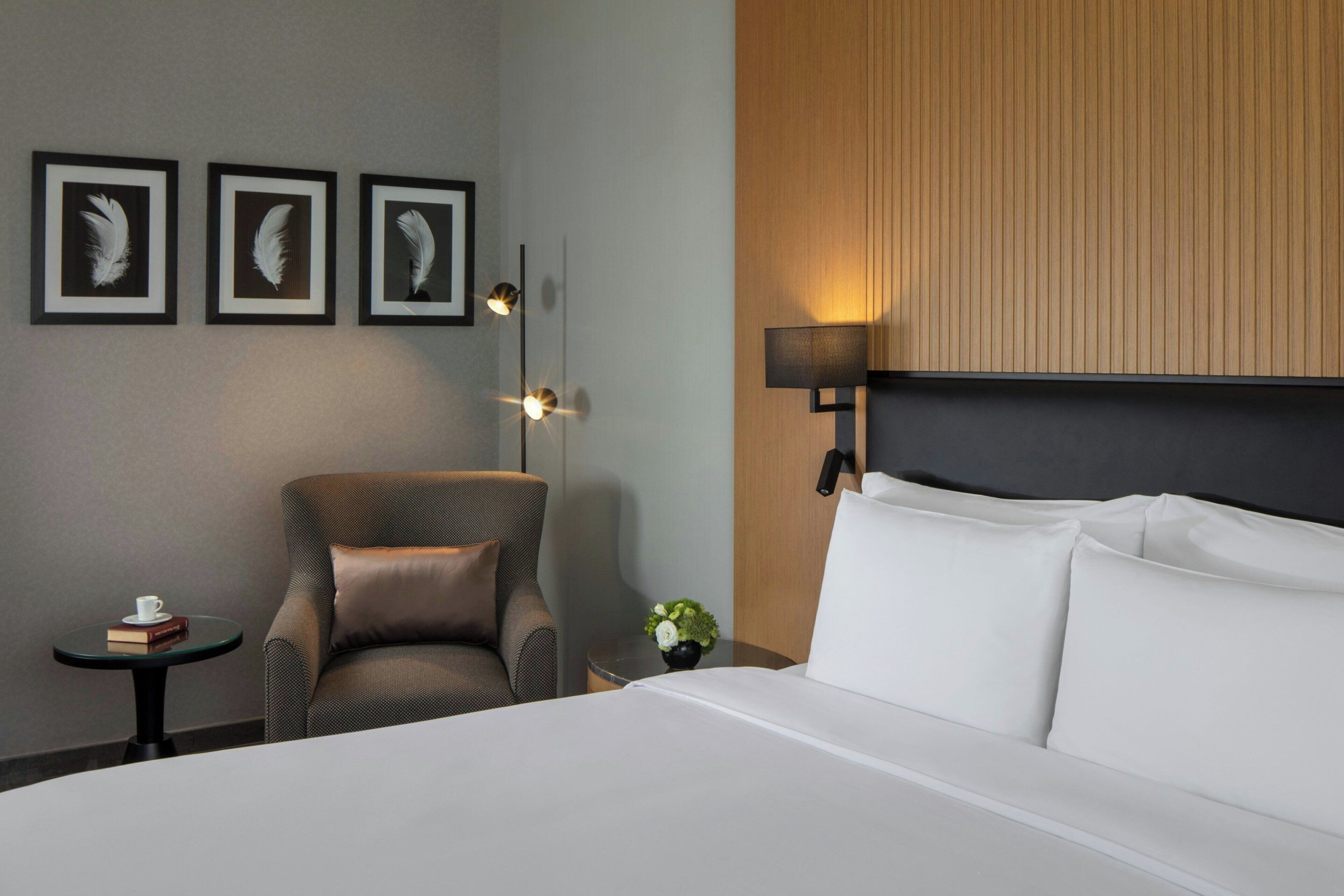 Quarto de hotel com cama, uma cadeira, uma mesinha com uma xícara e um livro, e três penas emolduradas na parede.