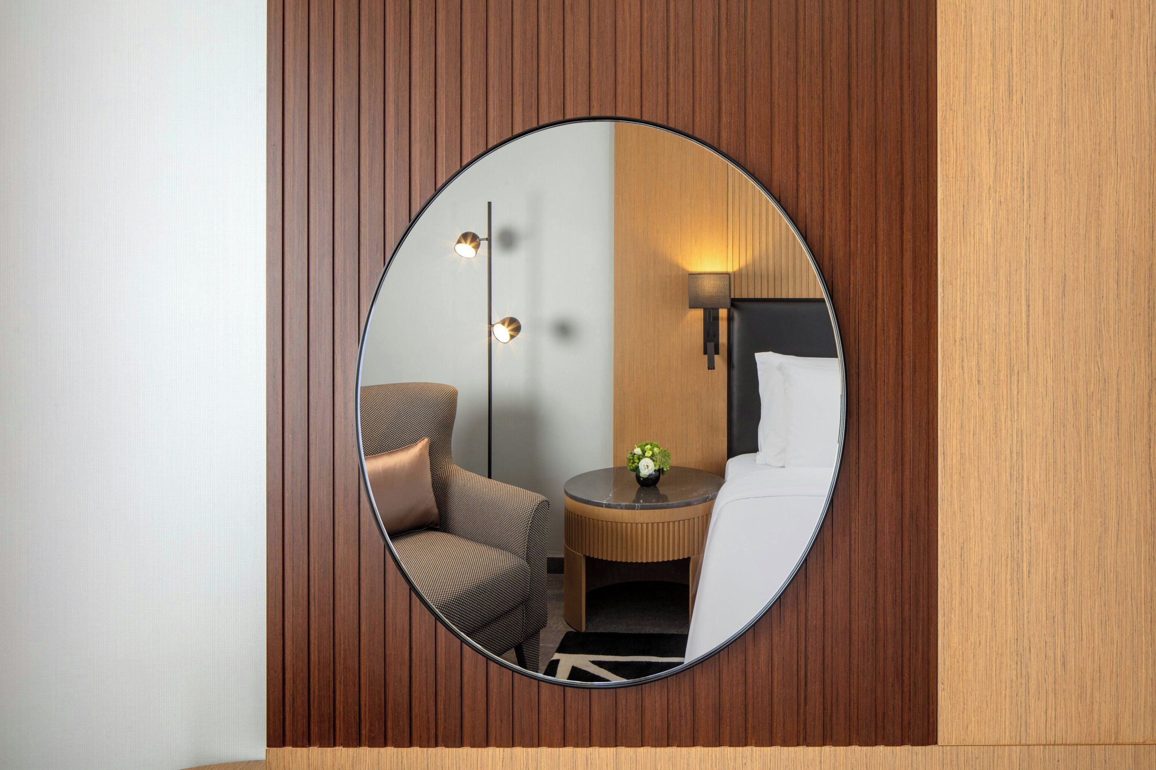 Un miroir rond reflétant une pièce dans laquelle on peut voir un lit, une chaise, une petite table avec un vase et une lampe.