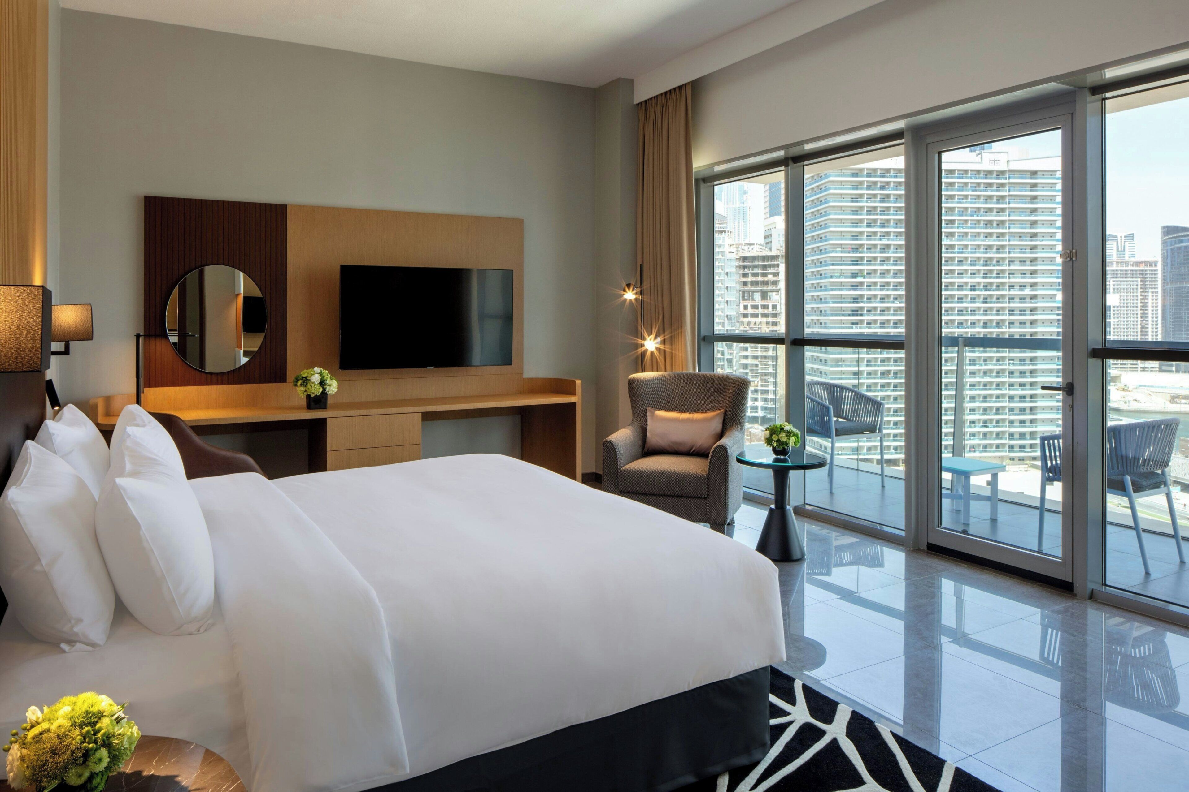 A hotel room with a large bed, a TV, a desk, a chair, and a balcony with a city view.