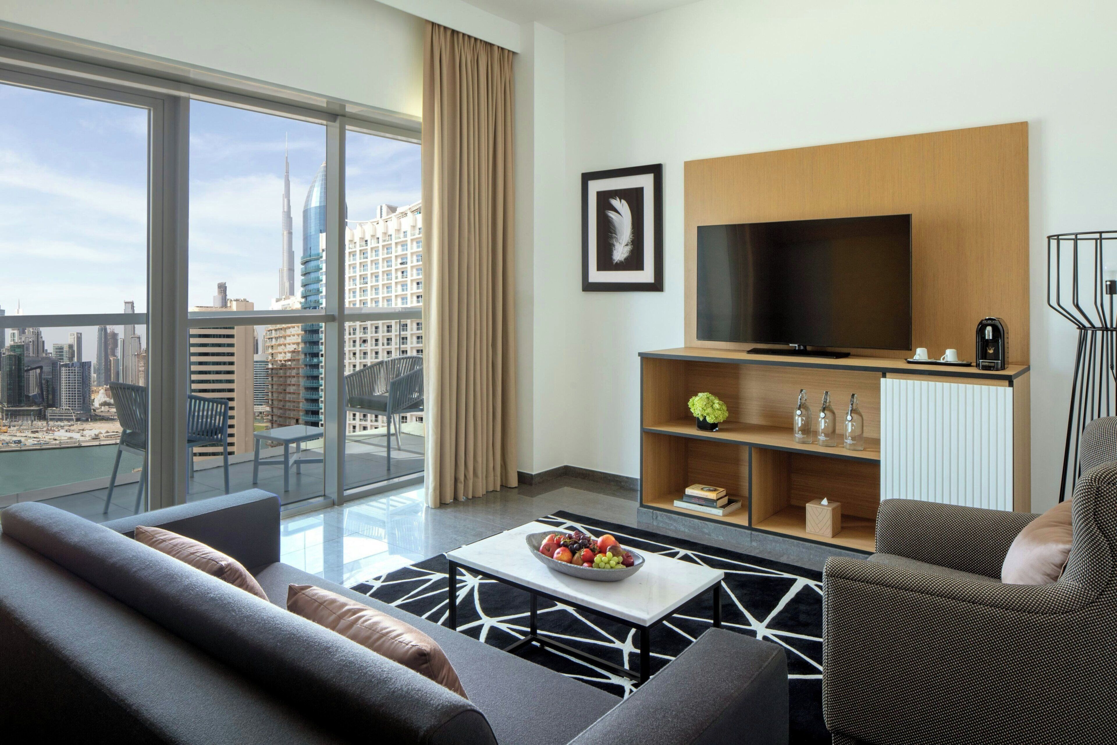 Une chambre d’hôtel moderne dotée d’une grande télévision, d’un canapé, d’une table basse avec des fruits et offrant une vue sur le panorama urbain.