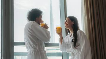 Duas pessoas vestidas de branco bebendo suco de laranja em uma mesa de café da manhã, com frutas e vista para um edifício.