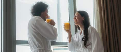 Two people in white robes drinking orange juice at a breakfast table with fruit and a view of a building.