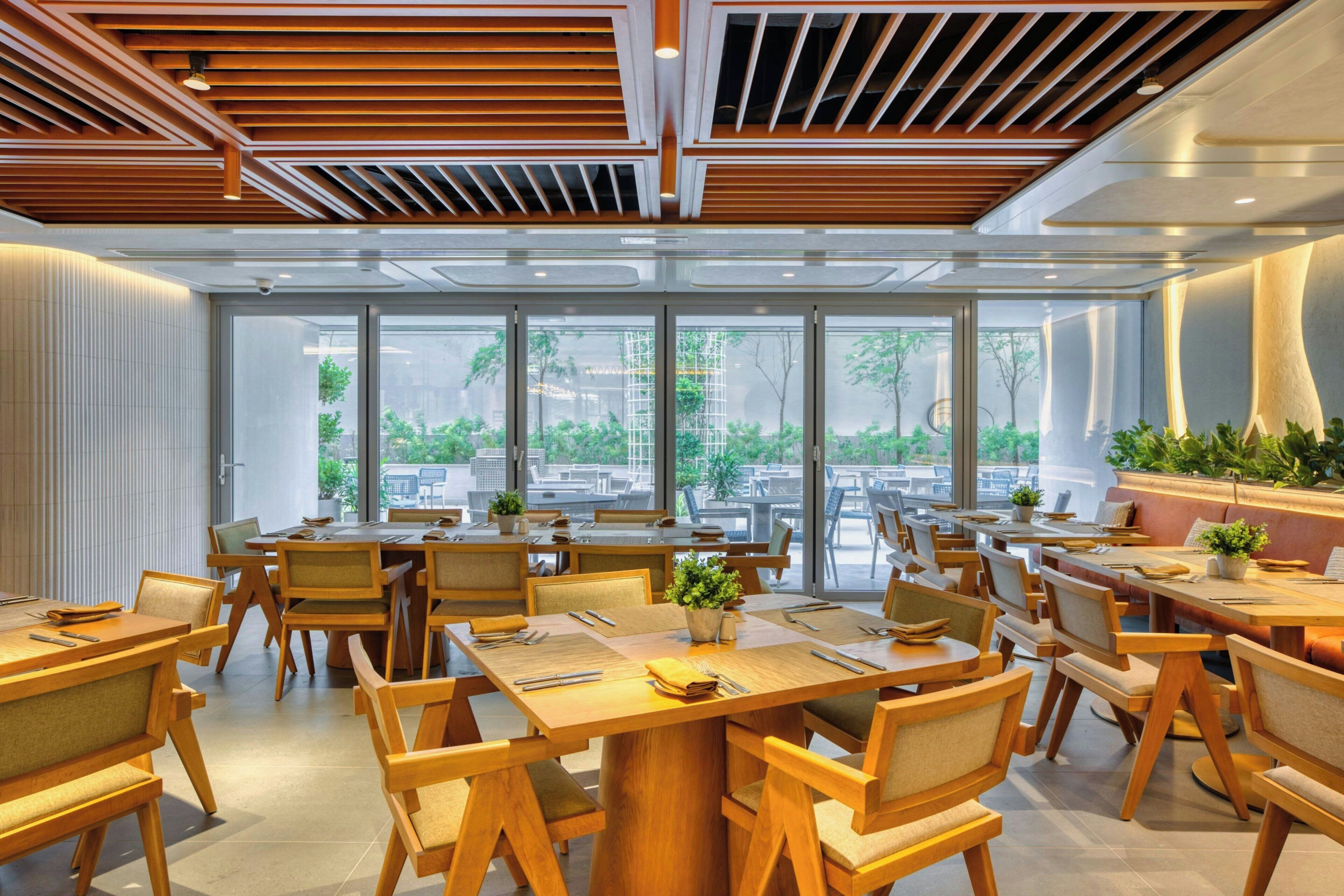 A modern restaurant with wooden furniture, large windows, and a view of greenery.