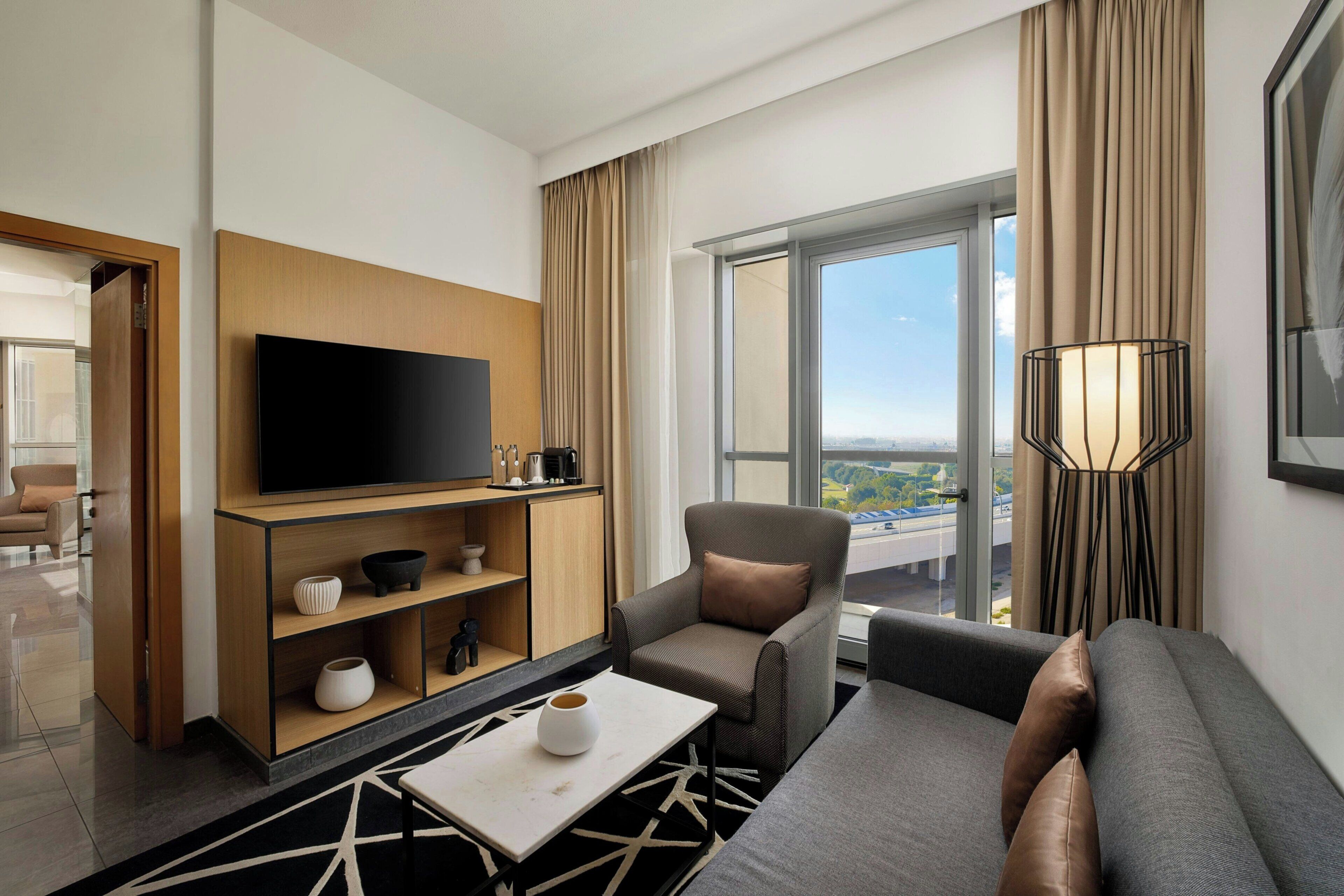 A modern hotel room with a flat-screen TV, a sofa, a chair, a coffee table, and a large window offering a city view.