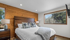Un dormitorio con cabecera de madera, un ventanal con vistas a árboles, una cama con ropa blanca y una mesita de noche con lámpara.