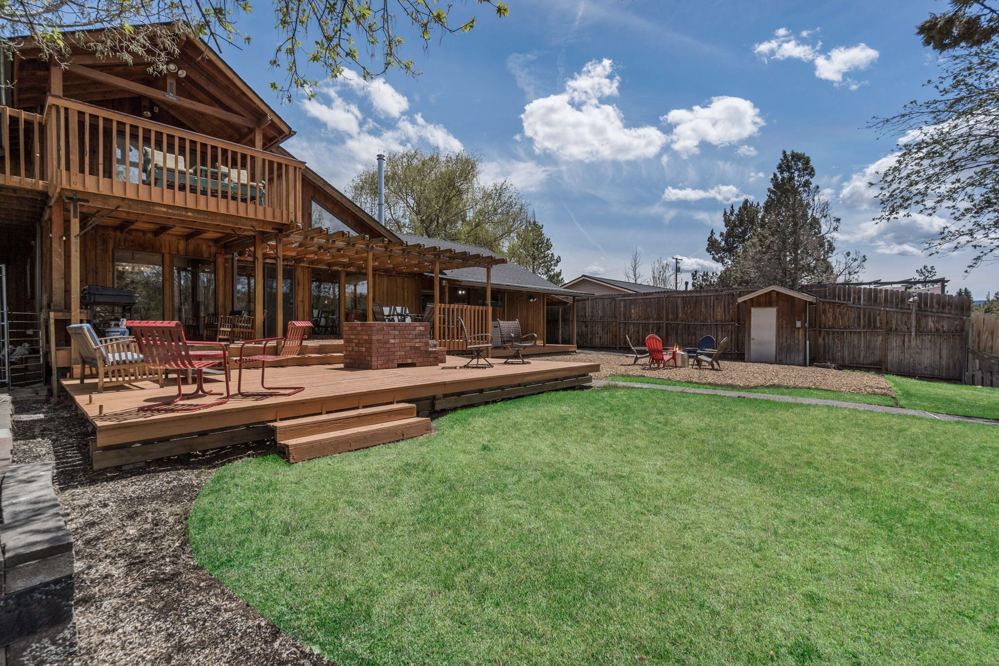 Una terraza de madera con mobiliario exterior, un porche cubierto y un jardín vallado.