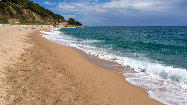 A sandy beach with gentle waves, a clear sky, and a rocky cliff in the background.