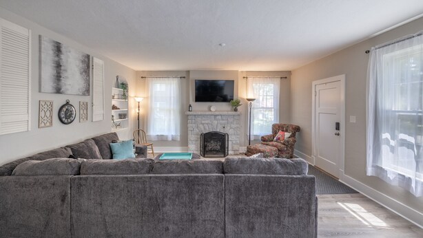 A living room with a gray sectional sofa, a fireplace, a flat-screen TV, and a large window with sheer curtains. - New Remodel in St. Charles - Sleeps 10! New patio! Large Fenced-in backyard! (Saint Charles)
