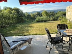A patio with a table and chairs, overlooking a grassy area with trees and a distant hill.