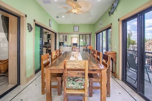 A dining area with a long wooden table and chairs, a ceiling fan, and large windows with a view of the outdoors.