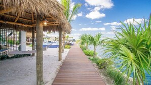 Un sendero de madera que lleva a una construcción con techo de paja, una piscina y palmeras.
