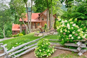 Une cabane rustique avec une cheminée en pierre, entourée d’une végétation luxuriante et d’une clôture en bois.