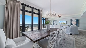 A spacious living room with a large wooden dining table, white wicker chairs, a grey sofa, and a balcony with a view of the beach. - New Rental!! Oasis, Formerly Phoenix West II, Lazy River and Slide!4th FLOOR! (Orange Beach)