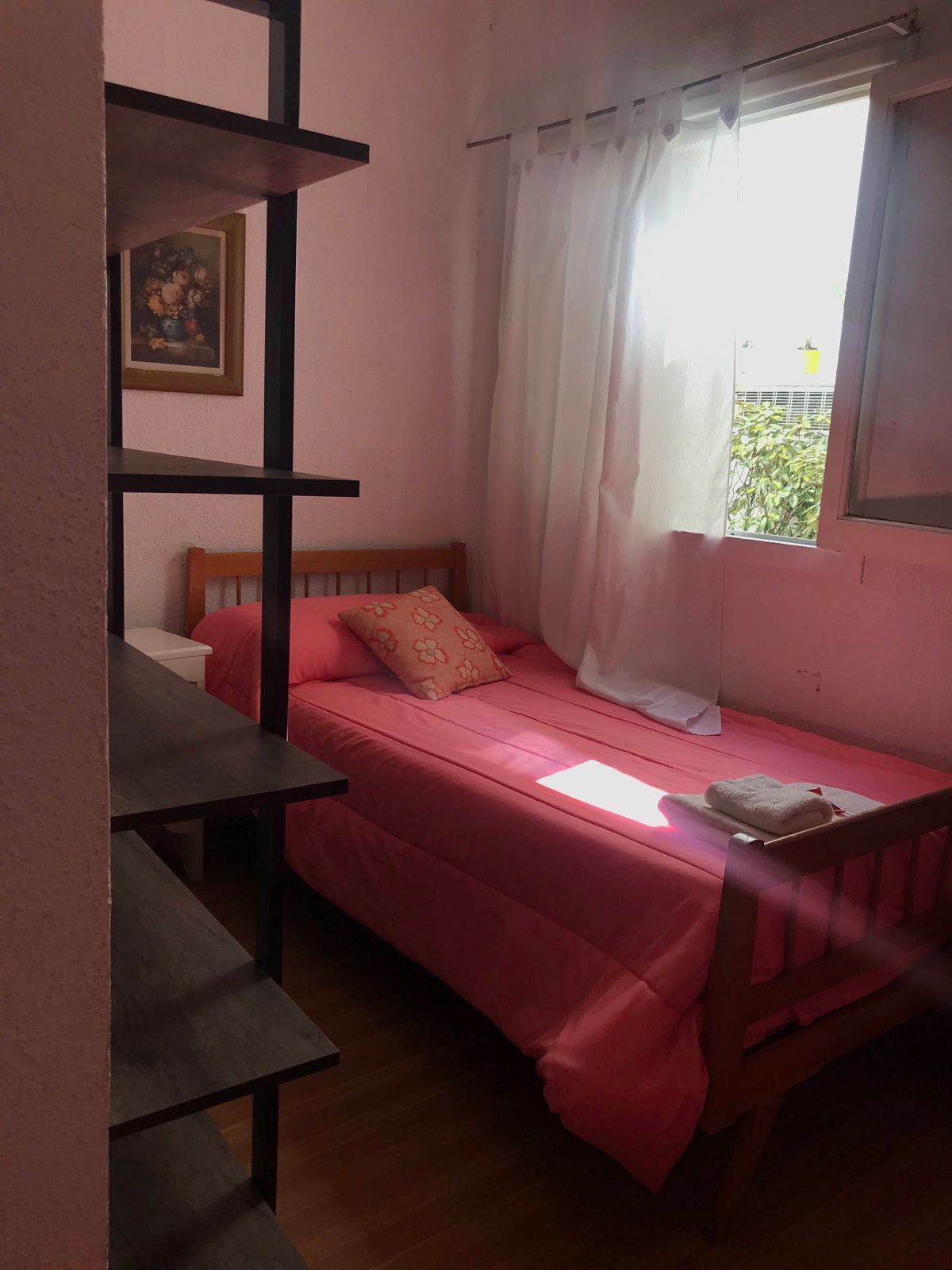 A small, single-bed room with a wooden headboard, a pink bedspread, a white pillow, and a window with white curtains.
