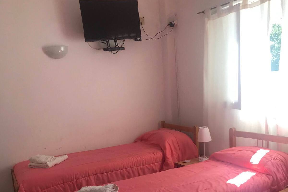 A room with two single beds, each with a pink bedspread and a white towel. There is a television mounted on the wall, a window with curtains, and a bedside table with a lamp.