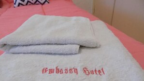 A white towel with "Embassy Hotel" embroidered in red on a bed with a pink bedspread. - Hotel Embassy (Miramar)