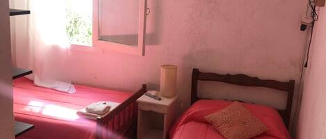 A room with two beds, each with a pink bedspread and a white towel. There is a small bedside table with a lamp and a framed picture on the wall.