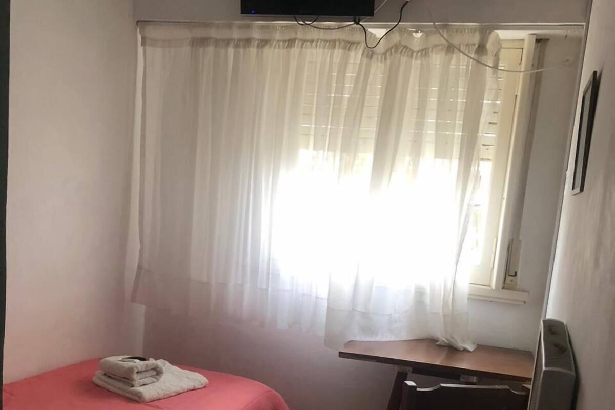 A small room with a bed, a desk, a chair, a television mounted on the wall, and a window with sheer curtains.