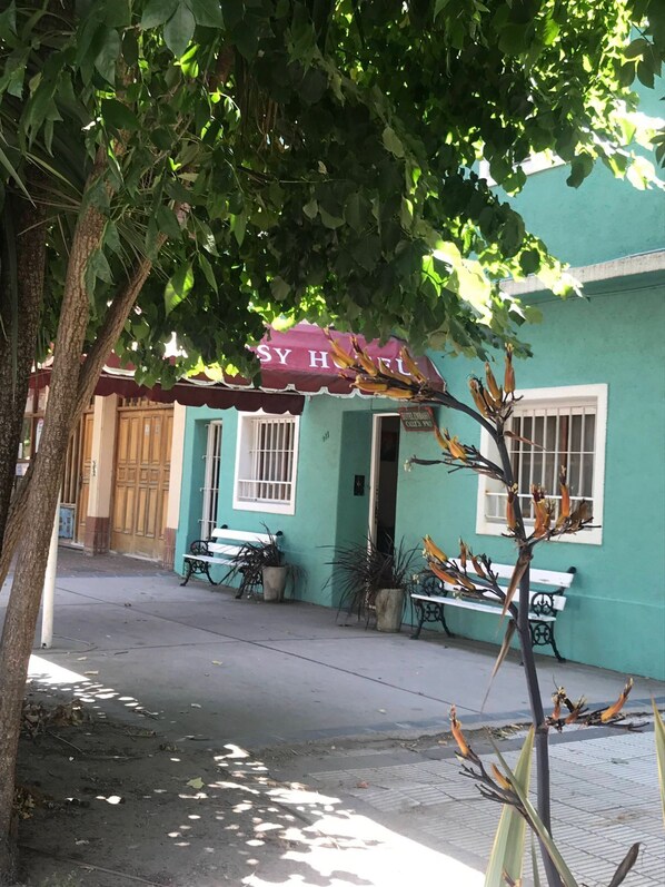 A hotel with a teal facade, a red awning, and a sign reading "SY HOTEL". There are potted plants and a bench outside.