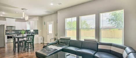 Una sala moderna con un sofá seccional, una mesa de centro de vidrio y un comedor, con una cocina al fondo.