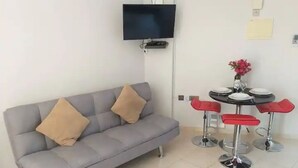 A living area with a grey sofa, two red bar stools, a flat-screen TV mounted on the wall, and a small table with a vase of flowers. - Panoramic Holidays - Deluxe 7 - Apartment with Outdoor Jacuzzi - Sea View (Peyia)