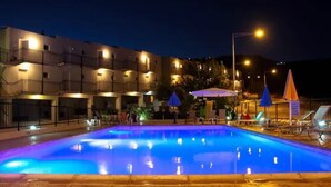 A swimming pool with a blue-lit edge, surrounded by lounge chairs and umbrellas, with a building in the background. - Panoramic Holidays - Deluxe 7 - Apartment with Outdoor Jacuzzi - Sea View (Peyia)