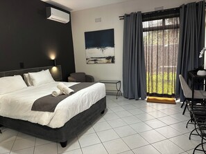 A modern hotel room with a large bed, a desk, and a chair. There is a window with curtains, a painting on the wall, and an air conditioning unit.