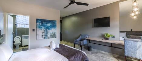 A modern hotel room with a bed, a desk, a chair, a ceiling fan, a TV, and artwork on the walls.