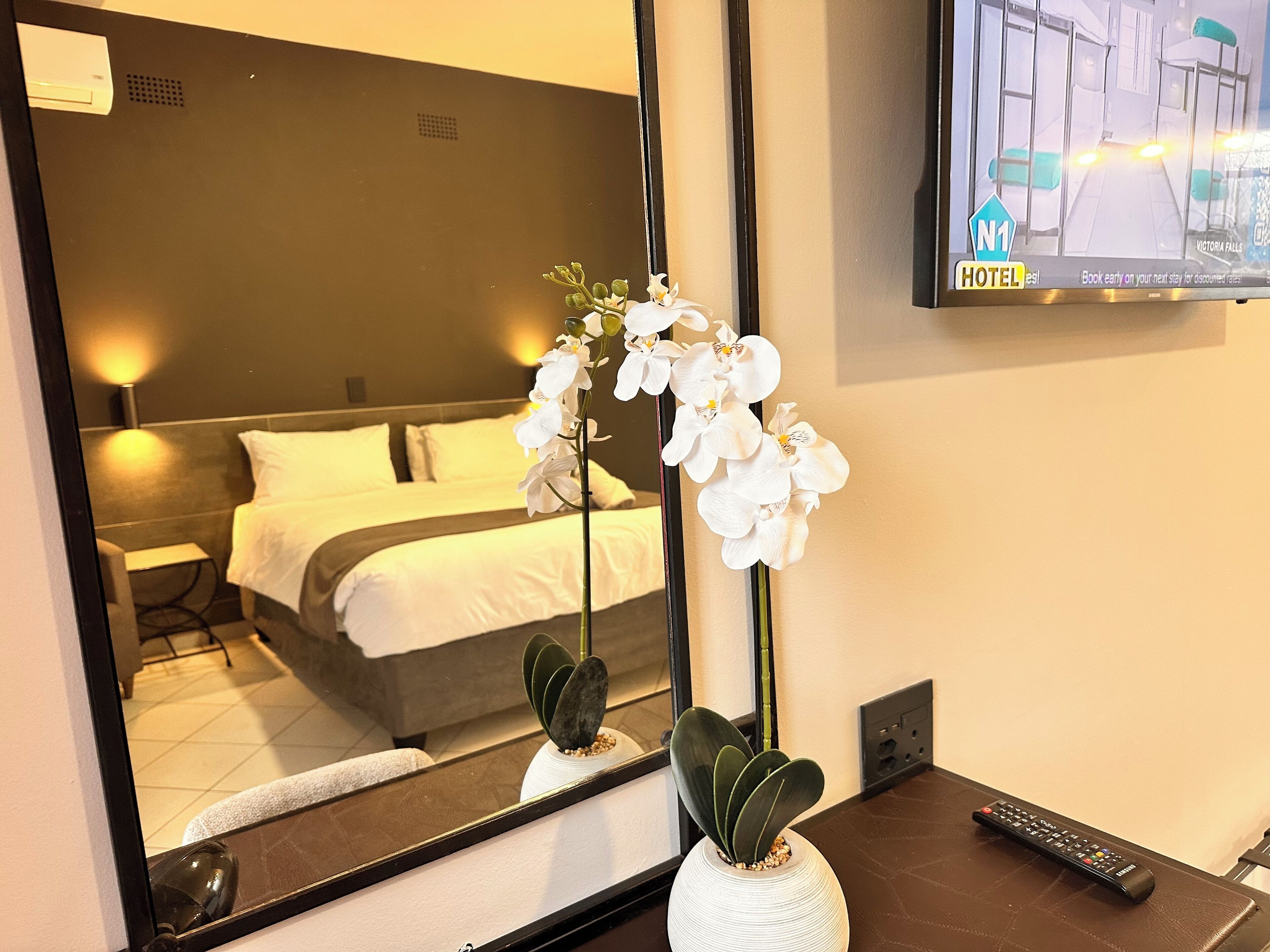 A hotel room with a bed, a mirror reflecting the room, a vase with white flowers, a remote control, and a TV displaying a hotel interior.