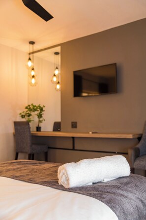 Moderni hotellihuone, jossa on siististi pedattu sänky, työpöytä tuolilla, seinälle asennettu televisio ja ruukussa oleva kasvi.