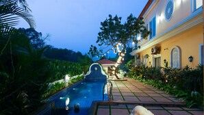 A swimming pool with a tiled deck, surrounded by lush greenery and a well-lit pathway leading to a building with a red roof. - Justa Casa Frangipani Assagao, Goa (Assagao)