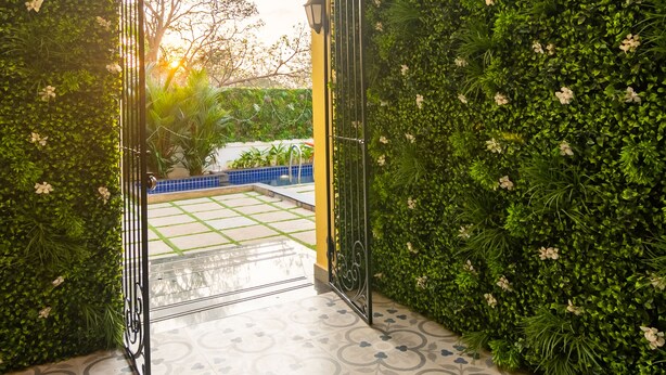 An open gate leading to a courtyard with a tiled floor and a green wall with white flowers. - Justa Casa Frangipani Assagao, Goa (Assagao)