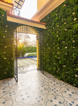 An open gate leading to a courtyard with a tiled floor and a green wall with white flowers. - Justa Casa Frangipani Assagao, Goa (Assagao)