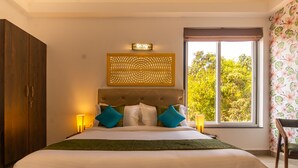 A bedroom with a large bed, a headboard with a decorative panel, two bedside lamps, and a window with a view of trees. - Justa Casa Frangipani Assagao, Goa (Assagao)
