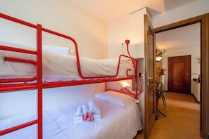 A bunk bed room with white bedding and red metal railings. There is a small table with a lamp and a chair.