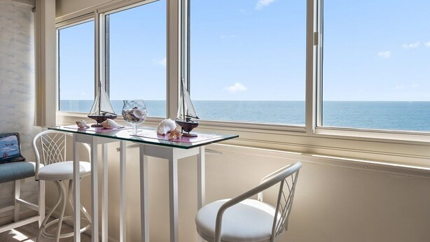 A room with a glass table and white chairs, a view of the sea through large windows, and a small sailboat model on the table.