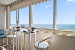 Une pièce avec une table en verre et des chaises blanches, une vue sur la mer par de grandes fenêtres et une petite maquette de voilier posée sur la table.