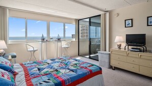 A hotel room with a large bed, a TV, a desk with chairs, and a balcony with a view of the sea.