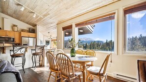 A cozy dining area with a wooden table and chairs, a view of mountains through large windows, and a kitchen with wooden cabinets and a microwave. - Snowflake Lookout: Ski In/Out Breckenridge Townhome with Hot Tub (Breckenridge)