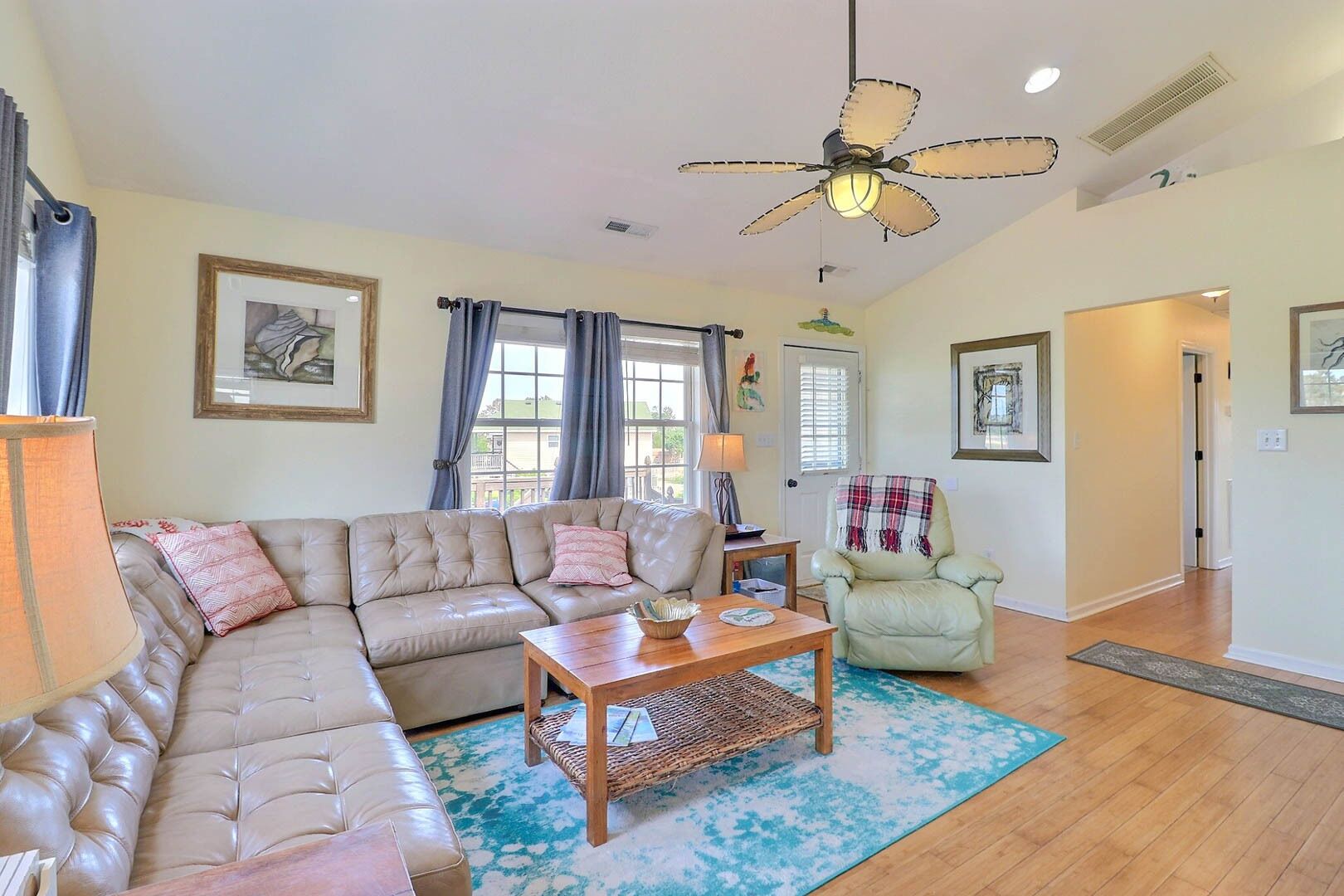 A living room with a sectional sofa, a wooden coffee table, a floor lamp, a ceiling fan, and a view of the outside through large windows.