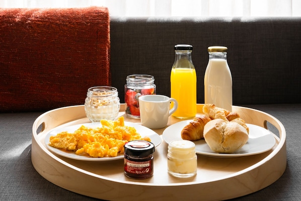 A breakfast tray with scrambled eggs, croissants, a glass of orange juice, a bottle of milk, a cup, and a jar of jam on a wooden tray.