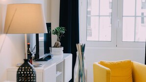 Un salón moderno con una consola blanca, un sofá amarillo, una planta en maceta y una ventana con vistas a edificios.