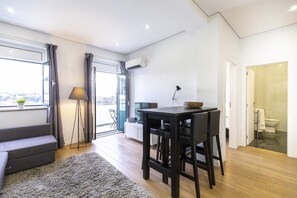 Una sala de estar moderna con un sofá, una mesa de comedor con sillas, una lámpara y vistas amplias por un ventanal.