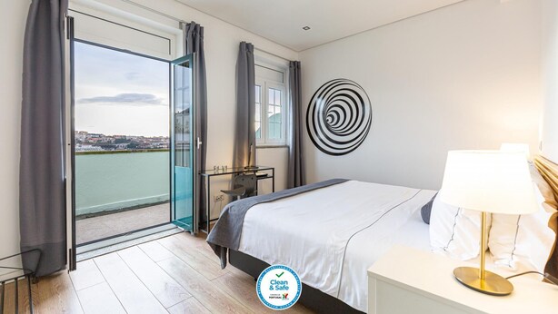 A modern bedroom with a large bed, a bedside table, a lamp, and a view of a river and cityscape through a glass door. - Feel Porto Historic Boutique Flat (Porto)