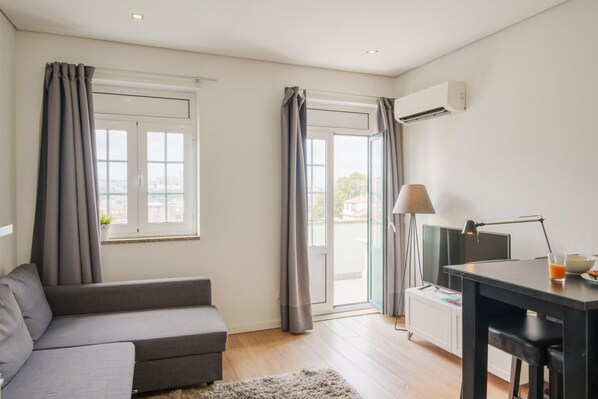 Una sala de estar moderna con un sofá gris, una mesita con una lámpara y un televisor. Hay dos ventanas con cortinas y un equipo de aire acondicionado en la pared.