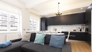 A modern living room with a dark grey sectional sofa, a chandelier, and a view of the kitchen with dark cabinets and white countertops. - Feel Porto Downtown Essence (Porto)