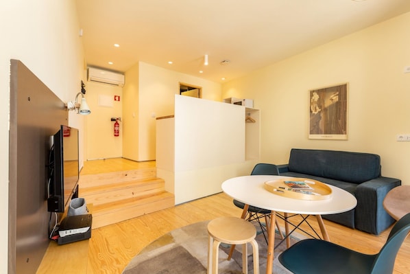 A modern living room with a sofa, a round table, and a television. There is a staircase leading to another room, and a fire extinguisher is mounted on the wall.