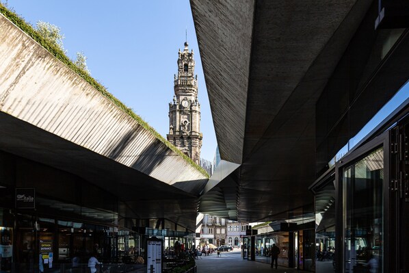 Un espacio peatonal urbano con una torre de reloj histórica al fondo, arquitectura moderna y un paseo techado.