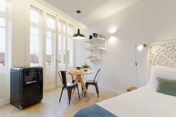 A modern room with a bed, a desk with chairs, a small table with a plant, and a vintage-style appliance.